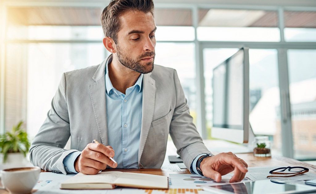 A man at a desk writing in a book while on his smart tablet. 